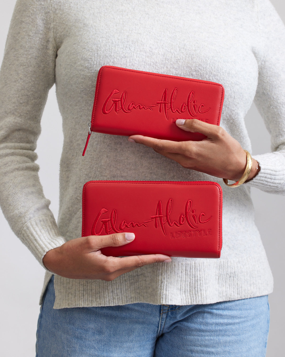 Red jumbo signature logo wallets with "Glam-Aholic" and "Lifestyle" embossed, held by a person in a beige sweater and jeans.