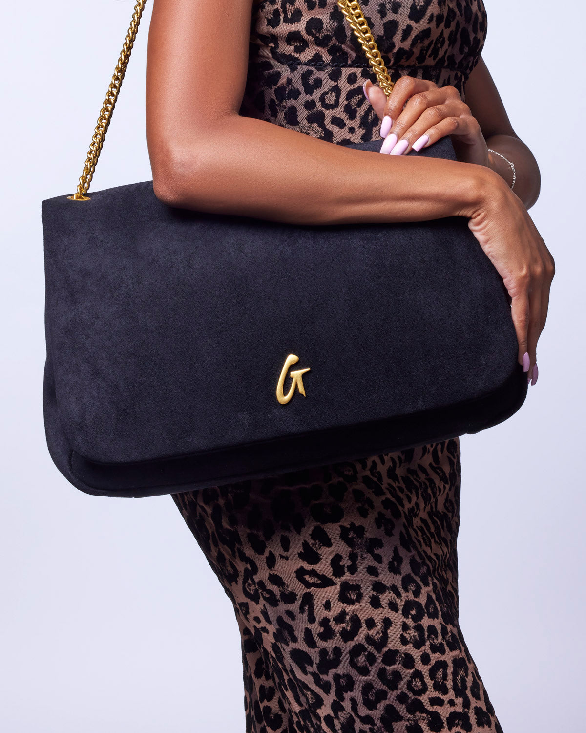 Large black suede flap bag with a gold logo, held by a model wearing a leopard print dress.