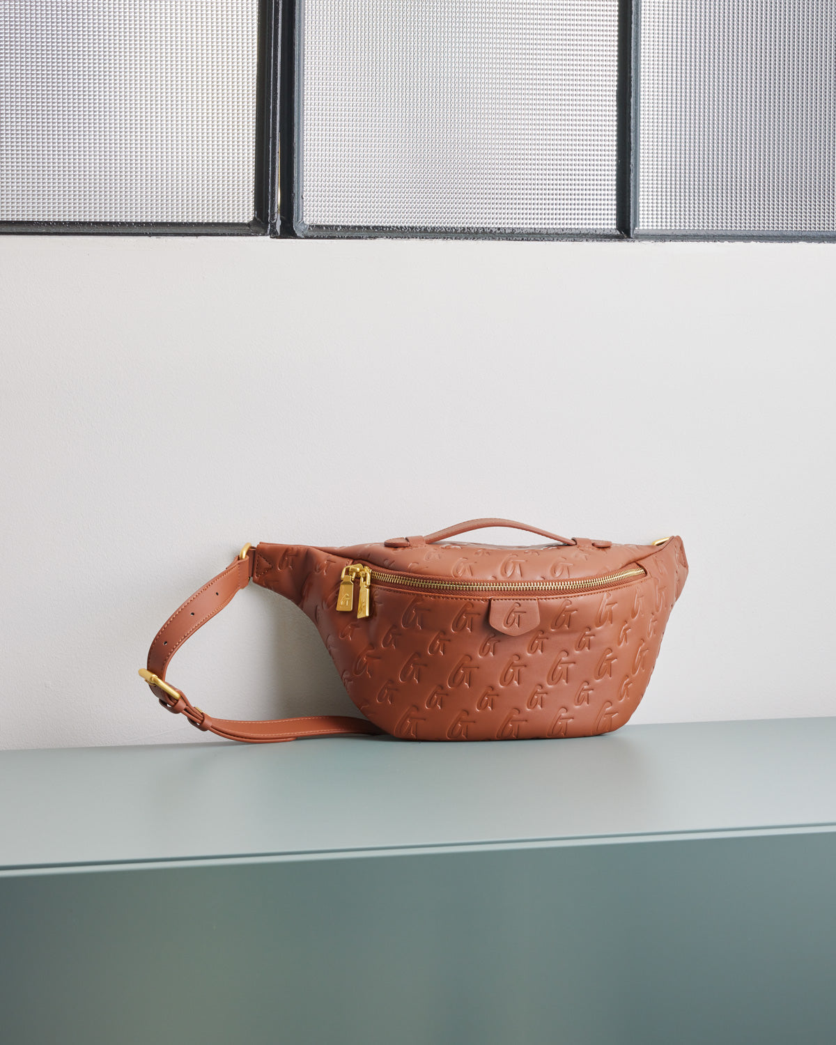 Brown monogram fanny pack with embossed logo pattern, gold zipper, and adjustable strap, resting on a light-colored surface.