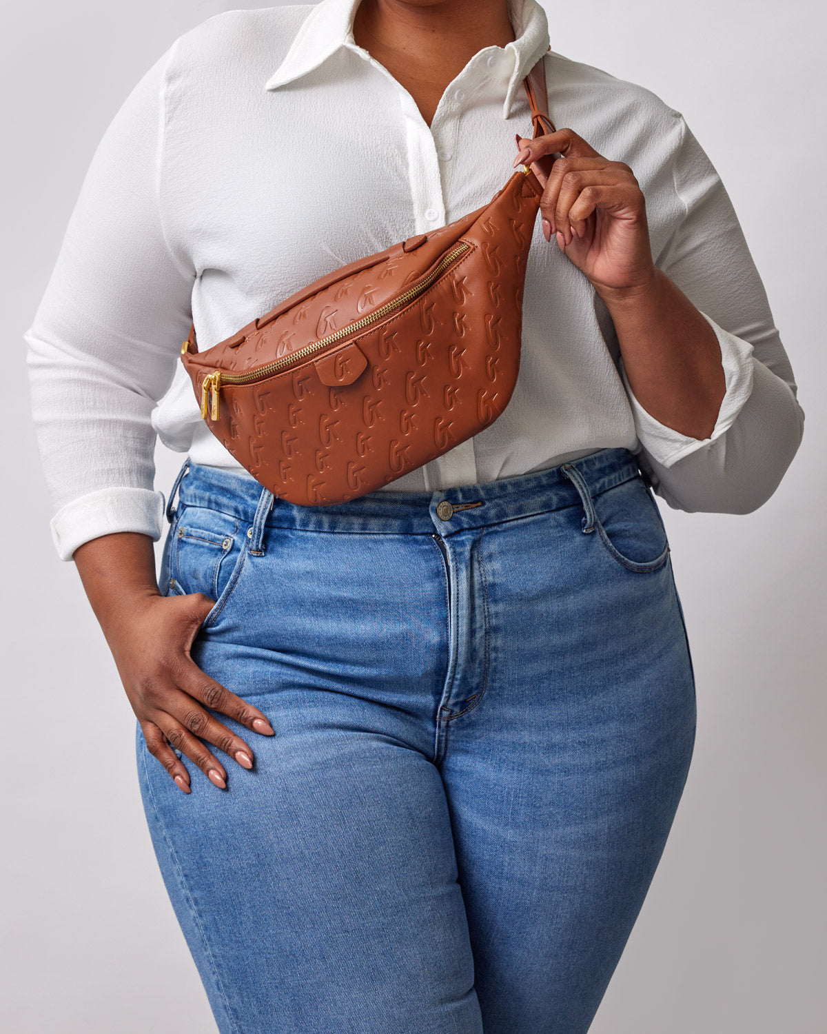 Brown monogram embossed fanny pack worn across the chest of a woman in a white shirt and blue jeans.