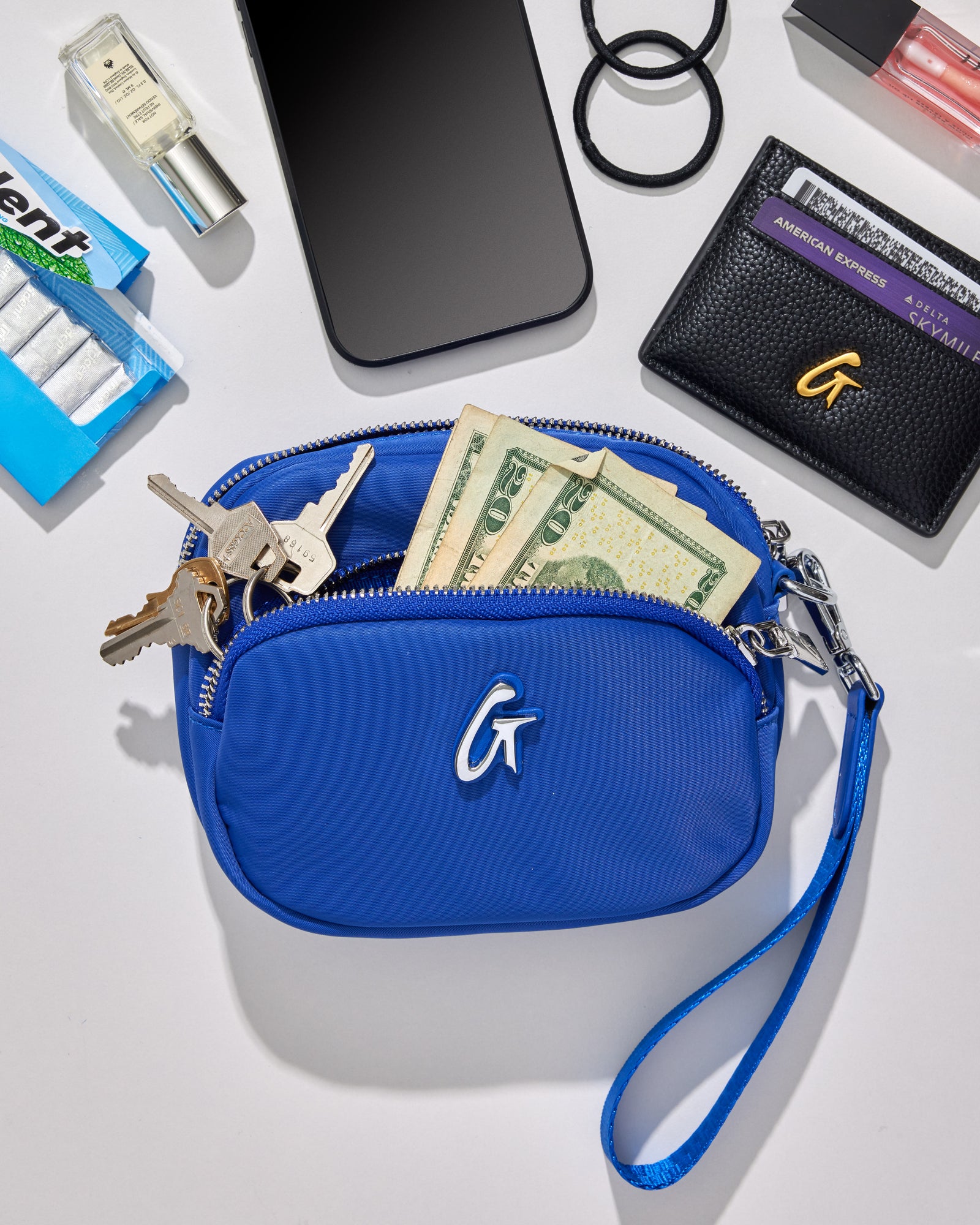 Blue nylon double zip wristlet with a wrist strap, partially open showing cash inside, surrounded by keys, phone, and accessories.