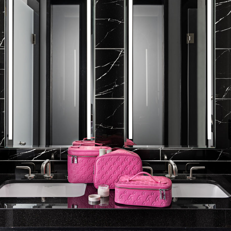 Large hot pink monogram toiletry bag set with embossed logo, placed on a black marble bathroom countertop near sinks.