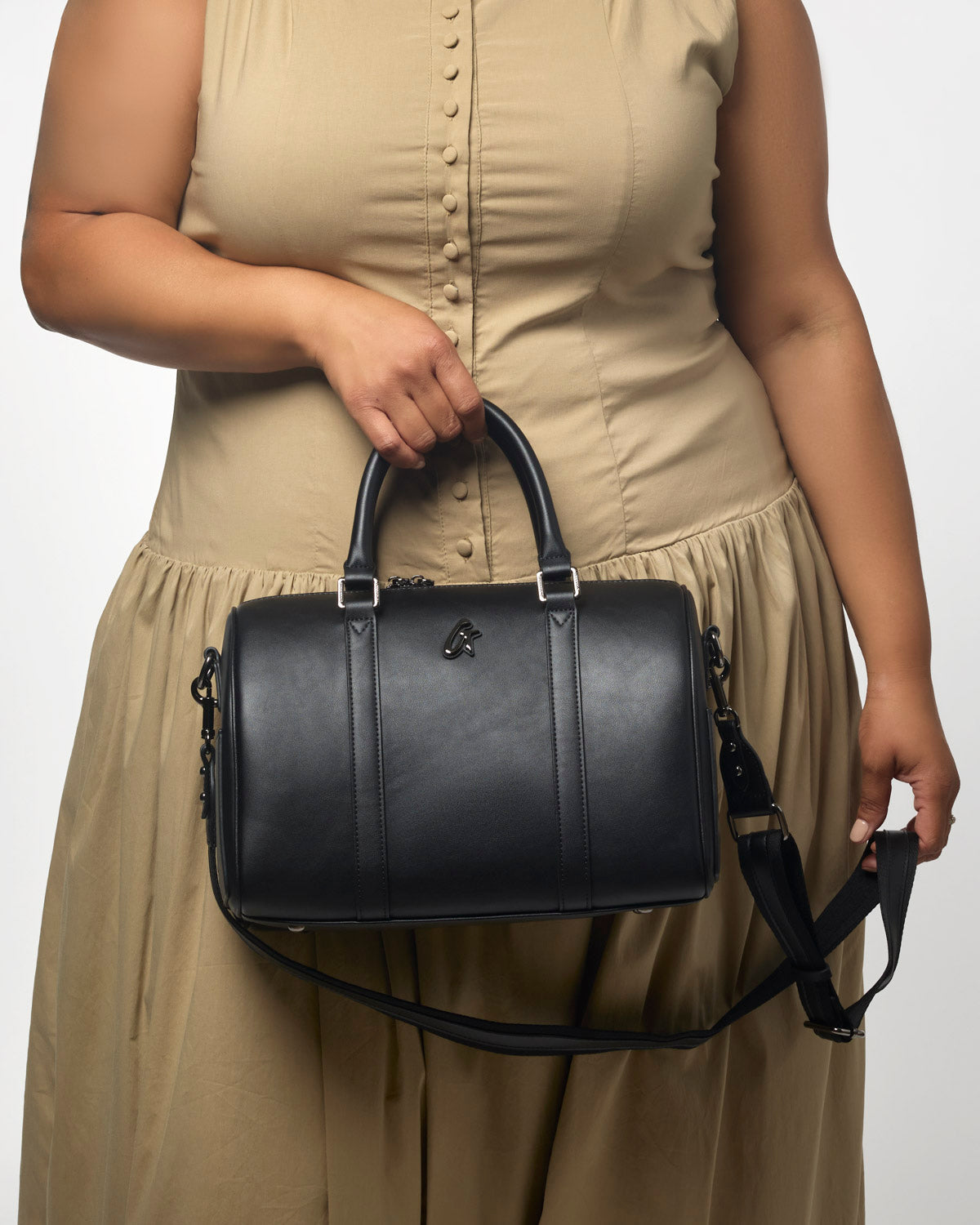 Medium black Boston bag with dual handles and a detachable shoulder strap, styled with a model in a beige dress.