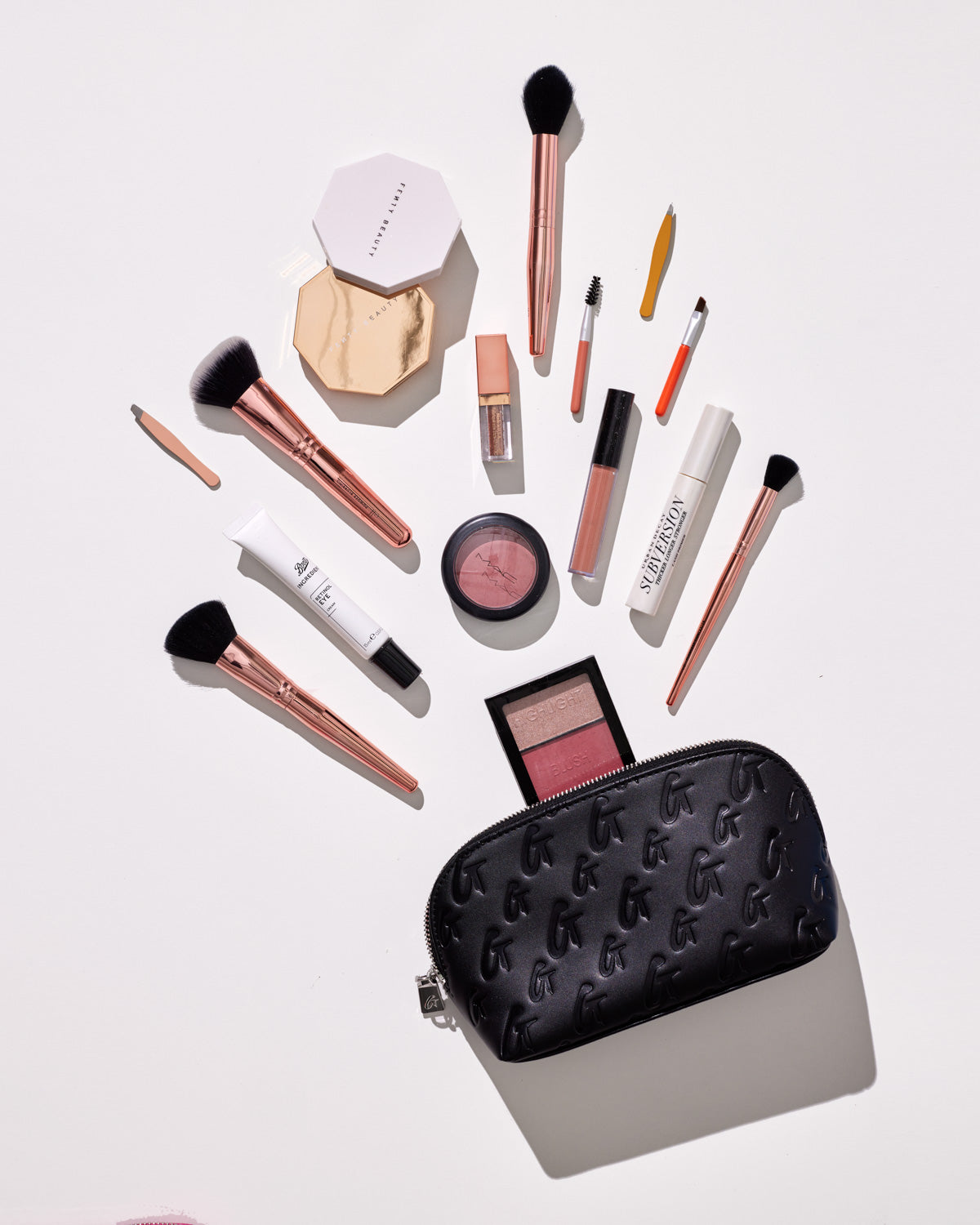 Black Monogram Cosmetic Pouch with embossed logo, surrounded by makeup brushes, lip gloss, blush, and beauty products on a white background.